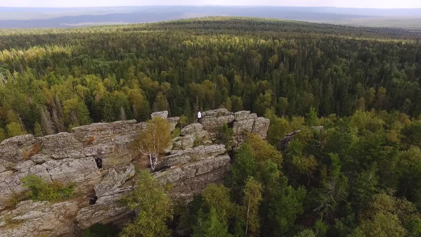 Beautiful high stone rock in green forest under blue sky. Footage. Top view of man standing on rock in thick green forest reaching horizon in autumn