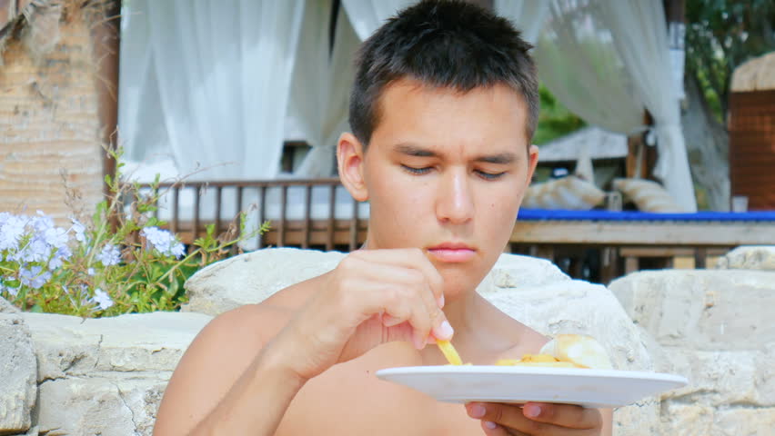 A young man with acne on his face eats french fries with ketchup, sitting in a cafe on the beach.