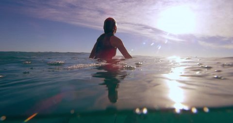 Female surfer sitting on surfboard in ocean in Australian, soaking up the sun waiting for the next big wave. Use for fitness/lifestyle advertising/commercial. Medium shot on 4k RED camera.