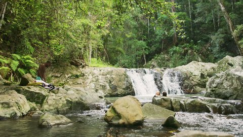 Four young, happy friends swim and dive in a clear pool in a stream with rocky banks with a waterfall in the background in an Australian forest. Wide shot in 4K on a RED Camera.