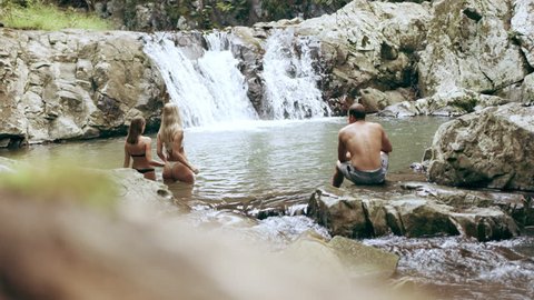 Two beautiful young women swim in a clear pool in a stream with rocky banks with a waterfall in the background as their friend watches, in an Australian forest. Wide shot, in 4K on a RED camera.