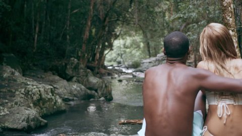 Young, fit, black man and his blonde caucasian girlfriend sit hugging on rocks overlooking stream in a forest. In natural sunlight, in Australia. Over the shoulder medium shot, in 4K on a RED camera.