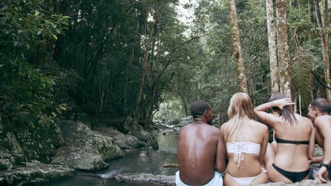 Four young friends sit in bathing suits and bikinis on rocks overlooking stream in a forest. In natural sunlight, in Australia. Over the shoulder medium shot, in 4K on a RED camera.