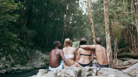 Four young friends sit in bathing suits and bikinis on rocks overlooking stream in a forest. In natural sunlight, in Australia. Over the shoulder medium shot, in 4K on a RED camera.