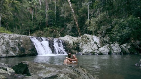 Young, happy, ethnically diverse couple swim and play in stream in front of waterfall while another friend dives in, in a forest in natural sunlight in Australia. Wide shot on 4K RED camera.