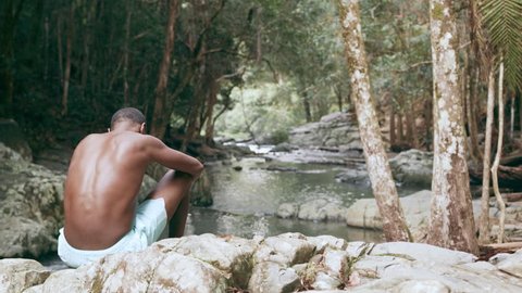 Young, fit, black man sits on rocks overlooking stream in a forest in natural sunlight, in Australia. Over the shoulder medium shot, in 4K on a RED camera.