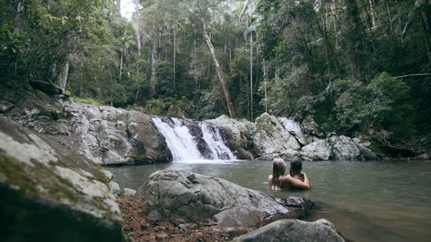 Young, happy, ethnically diverse couple swim and play in stream in front of waterfall in a forest in natural sunlight in Australia. Wide shot on 4K RED camera.