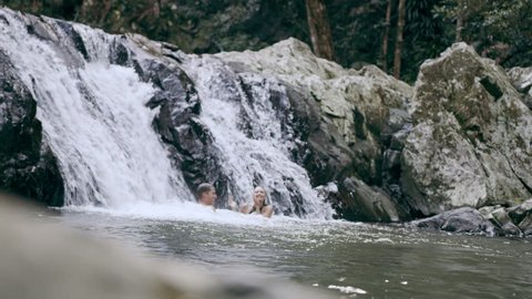 Young, happy, couple swim and play in stream in front of waterfall in a forest in natural sunlight in Australia. Wide shot on 4K RED camera.