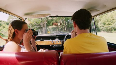 Young woman playfully photographs man as young couple drive a vintage car along a road in the country, natural daytime sunlight in Australia. Shot from the backseat, medium shot on 4K RED camera.