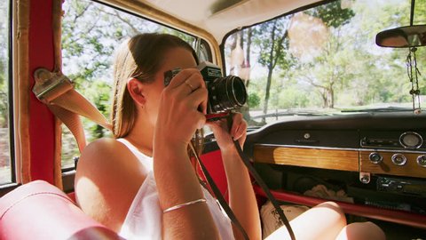 Enthusiastic woman photographs man as young couple drive a vintage car along a road in the country. Shot from the backseat, medium shot on 4K RED camera.