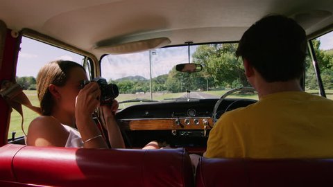 Playful woman photographs man as young couple drive a vintage car along a road in the country, natural daytime sunlight in Australia. Shot from the backseat, medium shot on 4K RED camera.