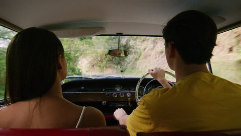 Happy woman puts phone to man's ear as young couple drive a vintage car along a road in the country, natural daytime sunlight in Australia. Shot from the backseat, medium shot on 4K RED camera.