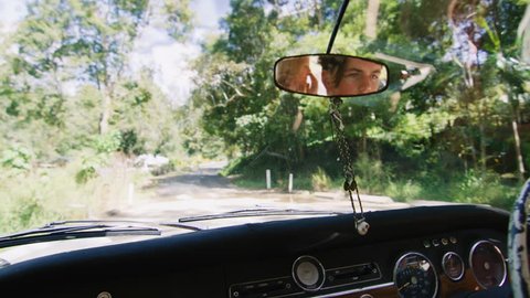 View from dashboard of vintage car, driving along country road Australian summer with happy young millennial man with hands on steering wheel. Shot in natural sunlight, medium shot on 4K RED camera.