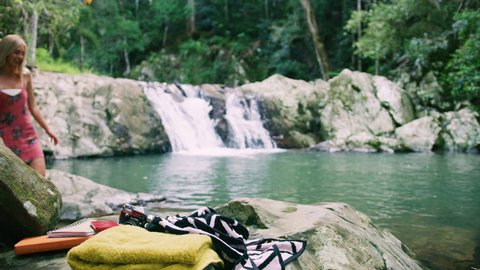 Two beautiful young women undress to bikinis to go swim in a clear pool in a stream with a waterfall in the background and with towels and camera in the foreground. Medium shot on 4k RED camera.