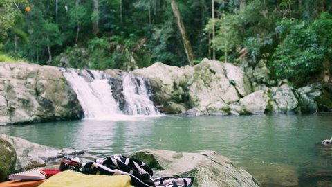 Two beautiful young women undress to bikinis to go swim in a clear pool in a stream with a waterfall in the background and with towels and camera in the foreground. Medium shot on 4k RED camera.
