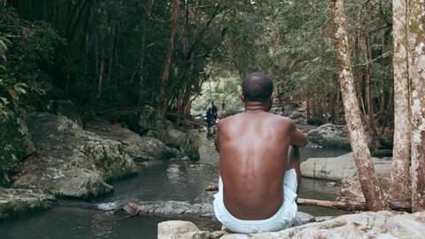 Young, fitman sits on rocks overlooking stream in forest when his blonde girlfriend sits next to him and holds his hand in Australia in natural sunlight. Over the shoulder medium shot on 4k RED camera
