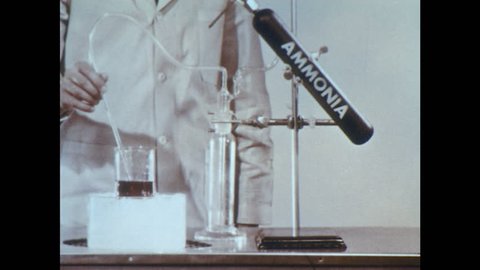 1960s: Lab. Man removes tubing from beaker and stops flow of ammonia. Man compares liquid in beaker to tray of test tubes. Man puts down beaker.