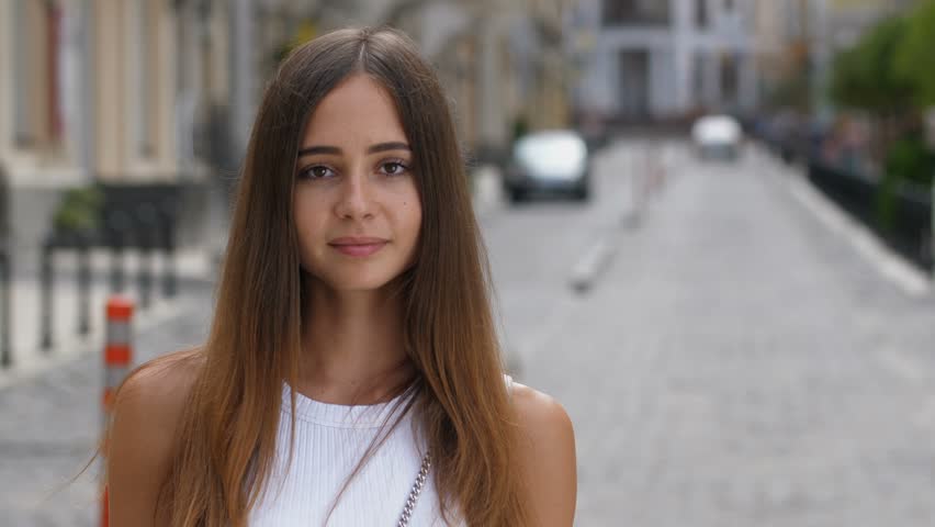Portrait of gorgeous caucasian girl with long brown straight hair and deep brown eyes smiling at camera outdoors. Joyful young woman with clean tanned skin looking at you with innocent enigmatic smile