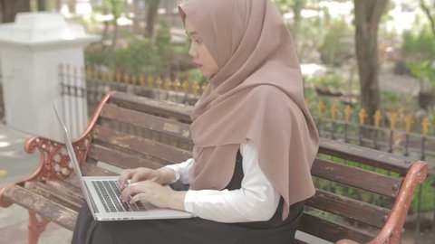 Young muslim woman typing / working on laptop. Woman with laptop sitting on bench at park.