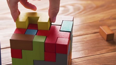 Hand folds colorful wooden cube puzzle on the brown table background, close up, dolly shot. Concept of decision making process, logical thinking. Geometric shapes on a wooden background.  