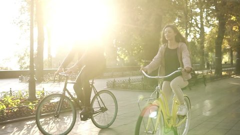 Close up of caucasian young couple or friends riding their bikes in the empty city park or boulevard in summertime. People, leisure and lifestyle concept. Green trees around, sun shines on the
