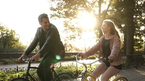 Caucasian young couple or friends riding their bikes in the empty city park or boulevard in summertime. Talking to each other, smiling. Sun shines on the background