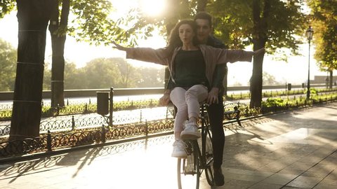 Young couple have fun riding on the same bike in outdoor activity with sun backlight on the background. Girl sitting on the rudder with outstretched hands. People, romance, leisure concept