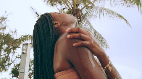 Beautiful black woman wearing peach bikini uses a shower on beach to wash off sand on a summer day with cloud cover in Australia. Medium shot in 4K on a RED camera.