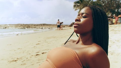 Attractive black woman wearing peach bikini lays on a towel on the beach and looks out to sea as people swim and surf in the background on an Australia summer day. Medium shot on 4K RED camera