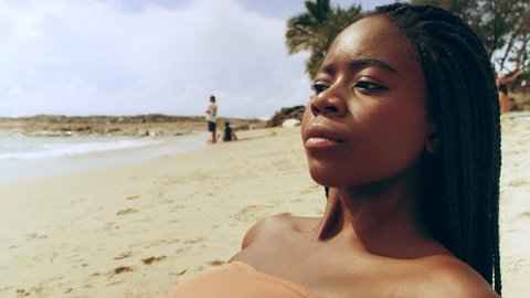 Attractive black woman wearing peach bikini lays on a towel on the beach and looks out to sea as people swim and surf in the background on an Australia summer day. Medium shot on 4K RED camera