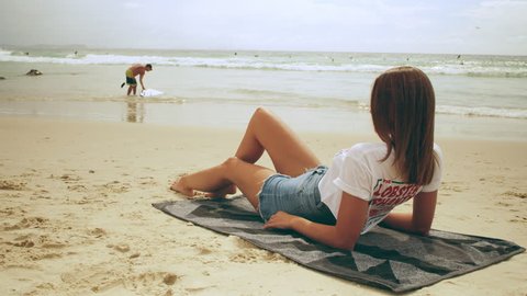 Beautiful young woman wearing cut off jean shorts and t-shirt lays on a towel on the beach on an Australian summer day. Medium shot on 4k RED camera.