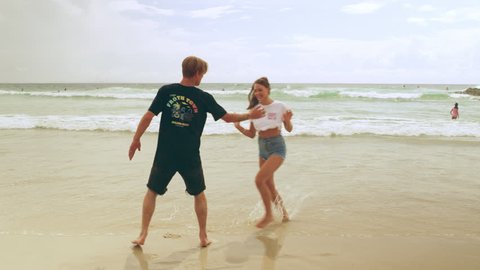 Happy, young couple joke and play on the beach as the waves lap around their feet on a summer day with clouds in the sky in Australia. Medium shot in 4K on a RED camera.