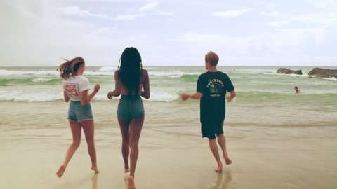 Three happy diverse, friends joke, splash and play in the surf on the beach as the waves lap around their feet on a summer day with clouds in the sky in Australia. Medium shot in 4K on a RED camera