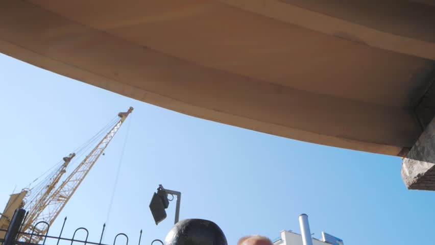 Young bearded male athlete training in industrial zone in sunny day, kettlebells exercises outdoors, urban background