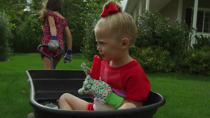 Tracking shot of girl pulling sister eating flavored ice in wagon in backyard / Pleasant Grove, Utah, United States