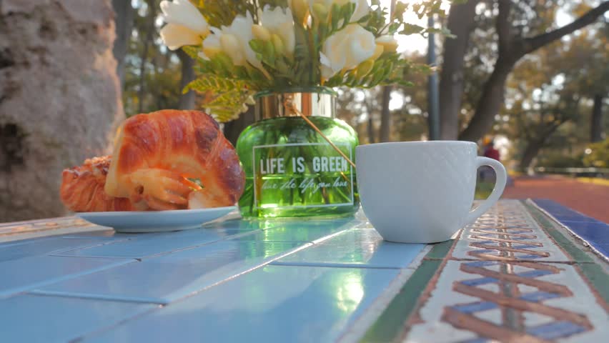 Two croissants, bouquet of white flowers, cups of tea in the park. Medialunas Argentinian sweet bakery. Green nature background and blue cozy table.