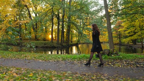 Woman go by ground path at autumn park, green and yellow leaves around, low evening sun flash through foliage. Lady walk at bank of small channel