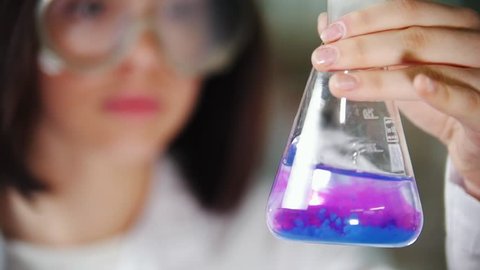Young woman in chemical laboratory holding a flask liquid and shaking it. The liquid changes color. Focus on the flask.