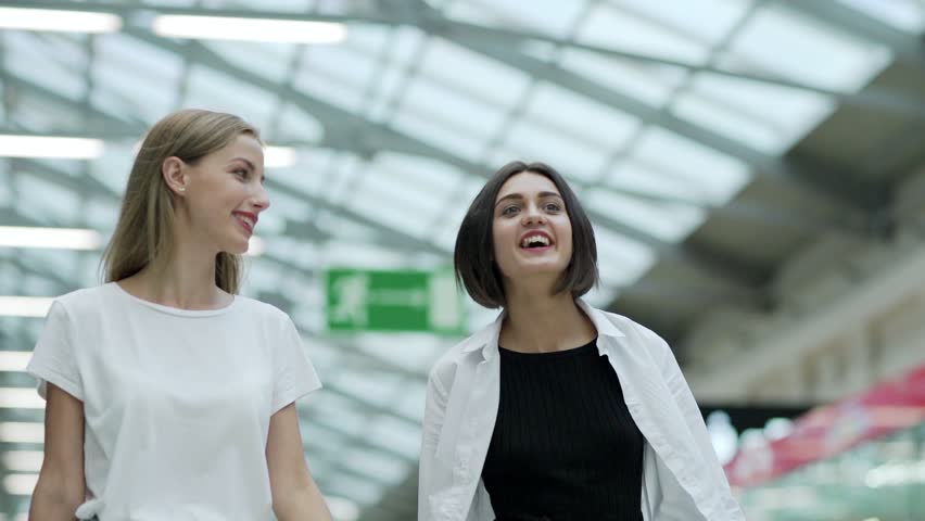 Dolly shot of two cheerful beautiful girls walking in shopping mall, talking and smiling