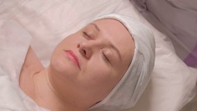 Middle-aged woman on the procedure in the cosmetology center. Close-up. The hands of a cosmetologist in lilac gloves put a soap solution on the female face. High quality raw video.