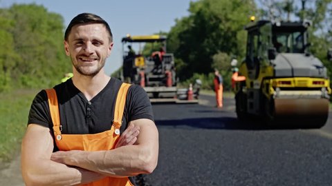 Portrait of a happy worker who repairs roads on the background of asphalt roller and other equipment. Happy employee looks at the camera.