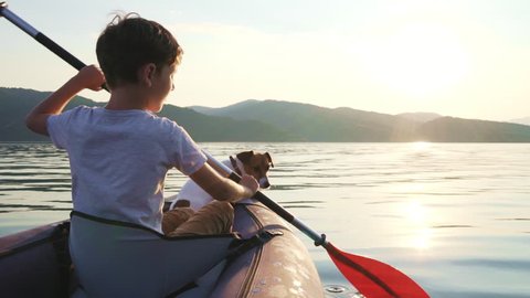 Happy boy with dog Jack Russell Terrier paddling an inflatable kayak on water of large mountain lake against backdrop of beautiful orange sunset slow motion. Family sports vacation. Tourism. Travel
