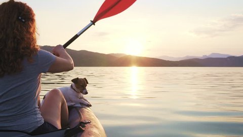 Happy young woman paddling on an inflatable kayak with her dog Jack Russell Terrier on water of large mountain lake against beautiful orange sunset slow motion. Family Sports. Summer. Travel. Tourism