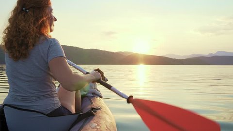 Happy young woman paddling on an inflatable kayak with her dog Jack Russell Terrier on water of large mountain lake beautiful orange sunset slow motion. Go Everywhere. Family Sports. Summer. Travel