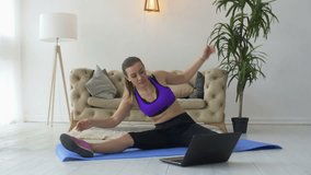 Lovely fit adult woman in sportswear doing distant training and stretching exercises with personal trainer using laptop pc while sitting on floor at home. Healthy lifestyle female exercising at home.