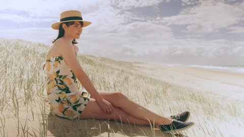 Beautiful brunette woman wearing a hat sitting on a beach looking out at the ocean in Australia with soft natural lighting. Wide shot on 4k RED camera.
