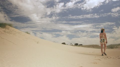 Beautiful brunette woman wearing a hat and dress walking along the sand of an Australian beach with soft natural lighting. Wide shot on 4k RED camera.