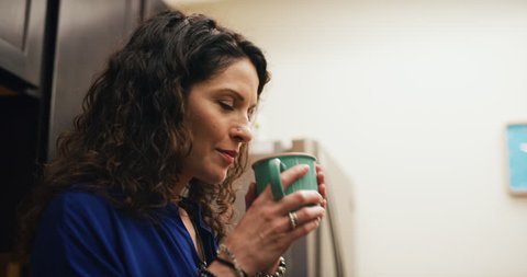 Content female office worker pours coffee smells and drinks it in office kitchen next to cake on a microwave. Medium to closeup shot on 4K RED camera on a gimbal.