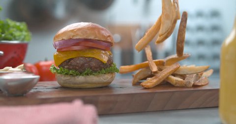 Delicious hamburger and salty fries falling onto a wooden board on a table with bright studio lighting. Close up slow motion shot on 4k phantom flex.