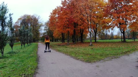 Woman slowly ride on self balancing board at autumn park, camera follow behind and move faster. Empty ground path at city park, old green grass and trees around, bright red leaves on maple trees